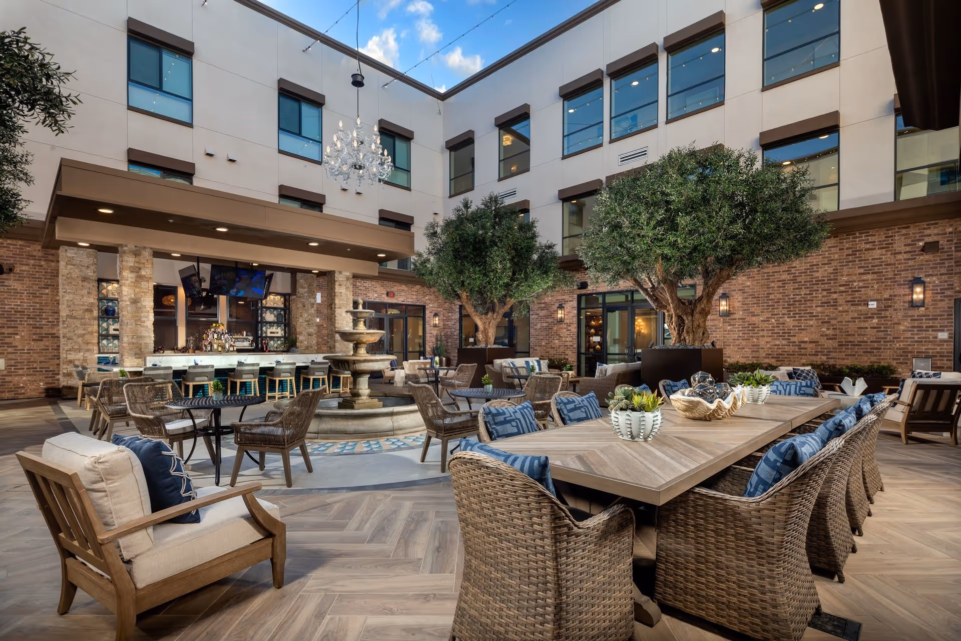 A furnished central courtyard with dining tables, wicker chairs, a fountain, a bar area, and potted trees beneath an open sky inside a multi-story building.