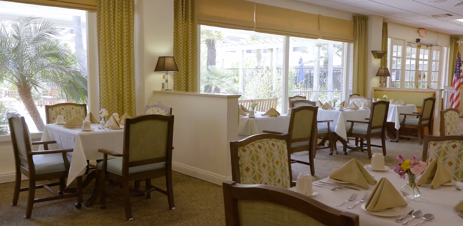 Bright dining room with tables set with folded napkins and chairs overlooking a garden through large windows.