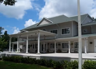 Exterior view of a large two-story building with a covered porch supported by white columns, surrounded by greenery under a partly cloudy sky.