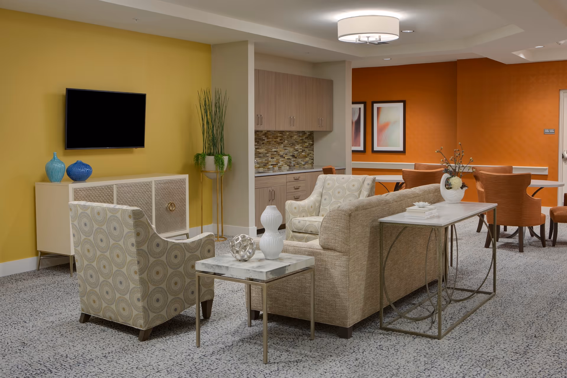 Bright communal living area with sofas and armchairs, a wall-mounted TV, and a dining table in the background.