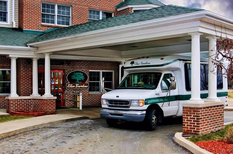 Entrance of a brick building with a covered driveway where a white and green shuttle bus labeled 'Elan Gardens' is parked. A sign with the name 'Elan Gardens' and a tree logo is visible near the entrance.