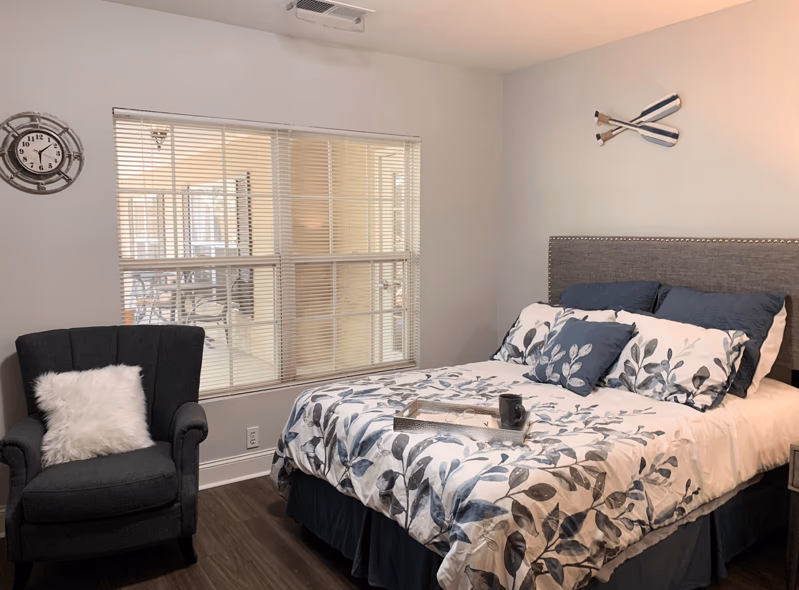 A cozy bedroom with a bed featuring a floral patterned comforter and multiple pillows. A dark upholstered armchair with a white fluffy pillow is positioned next to a window with blinds. A decorative clock is mounted on the wall to the left, and two crossed oars are hung on the wall above the bed's headboard.