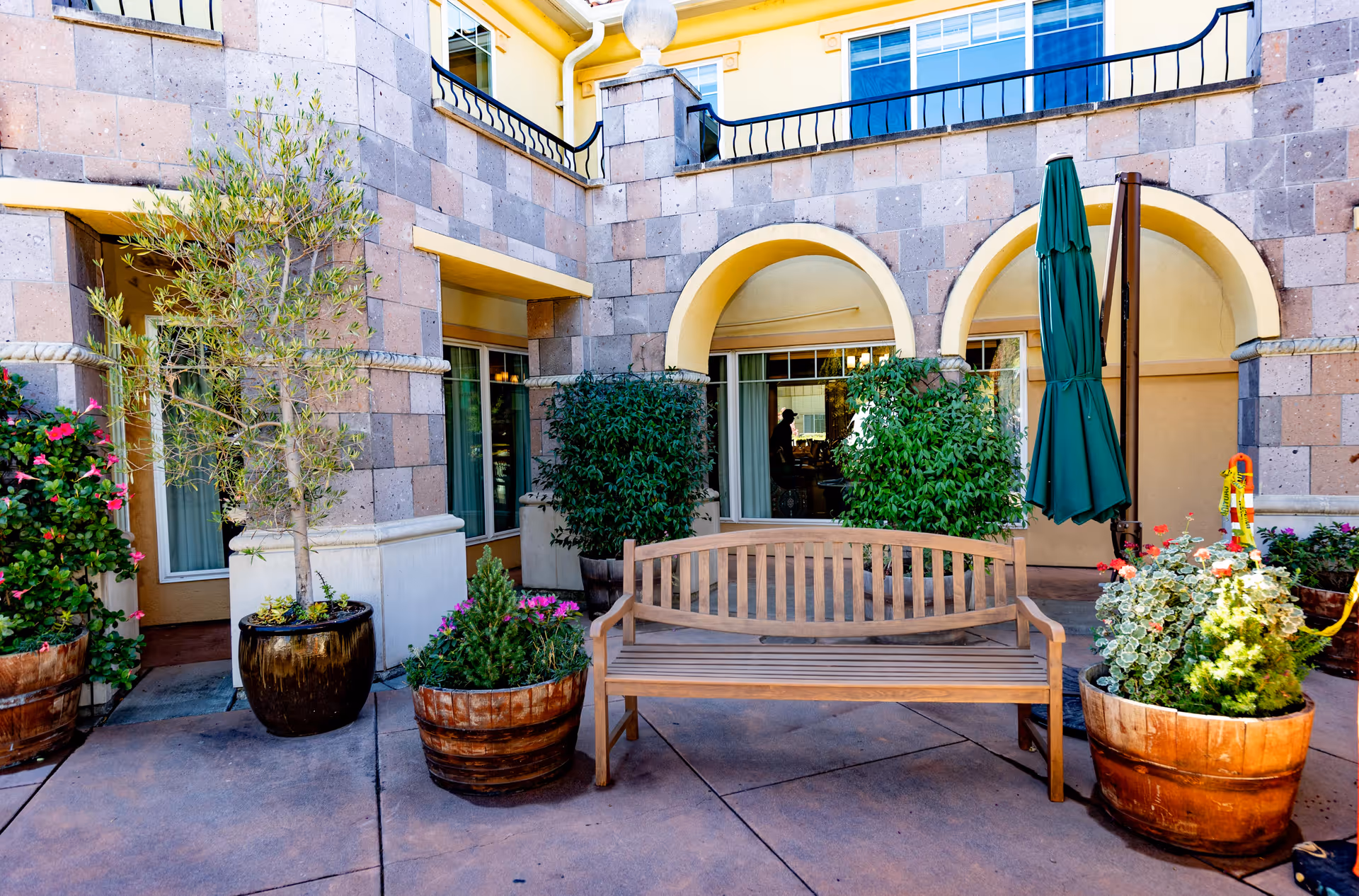 Courtyard with a wooden bench, potted plants, and a closed green umbrella in front of an arched building facade.