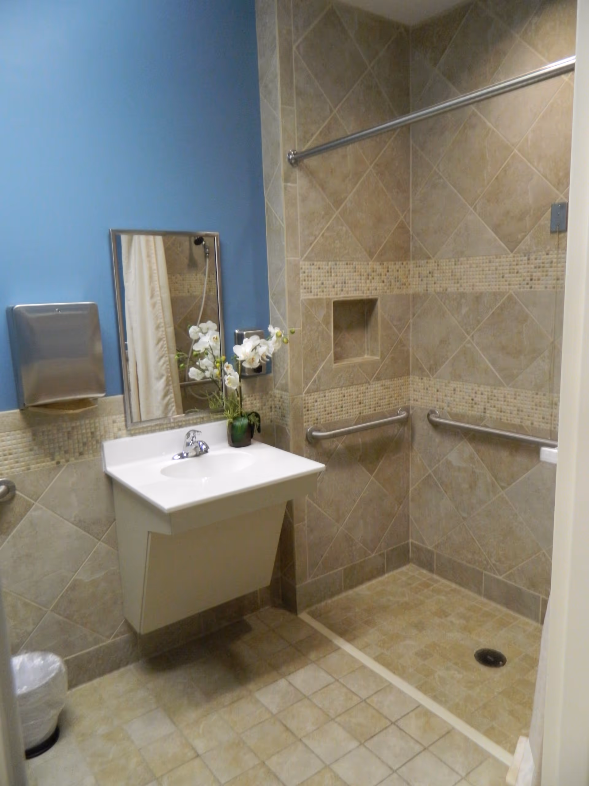 Accessible tiled bathroom with a wall-mounted sink, mirror, paper towel dispenser, and a roll-in shower with grab bars.