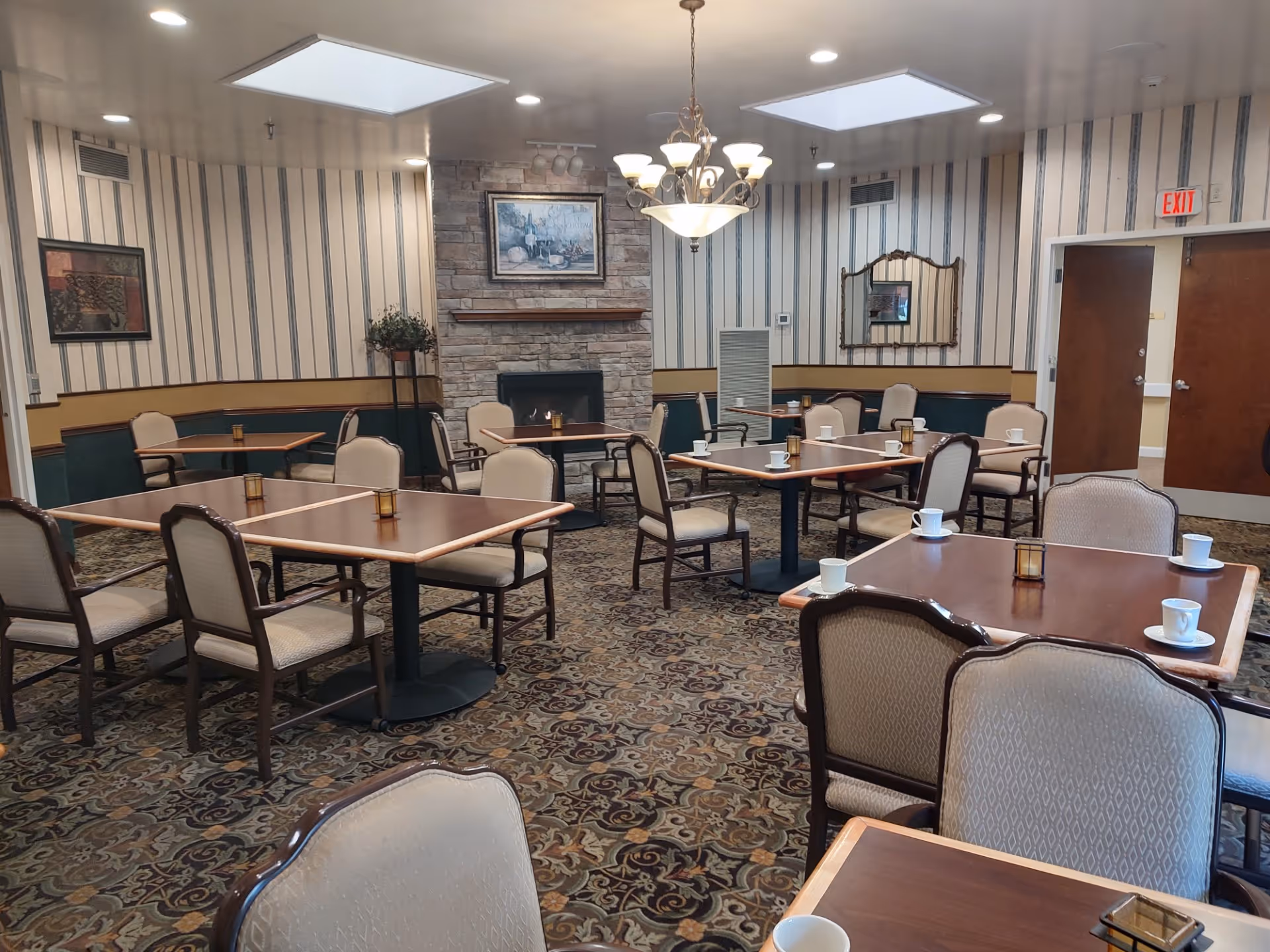 Dining room with multiple tables and chairs, a fireplace, chandelier, and cups set on the tables.