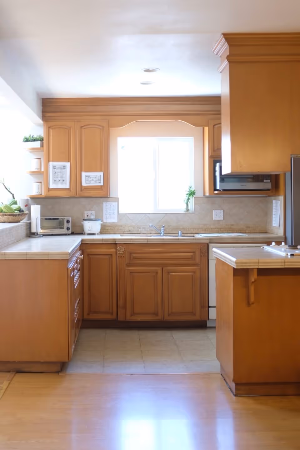 A bright kitchen with wooden cabinets and beige tiled countertops. There is a window above the sink letting in natural light. Kitchen appliances such as a microwave, toaster oven, and rice cooker are visible on the counters. The floor is a combination of wood and tile.