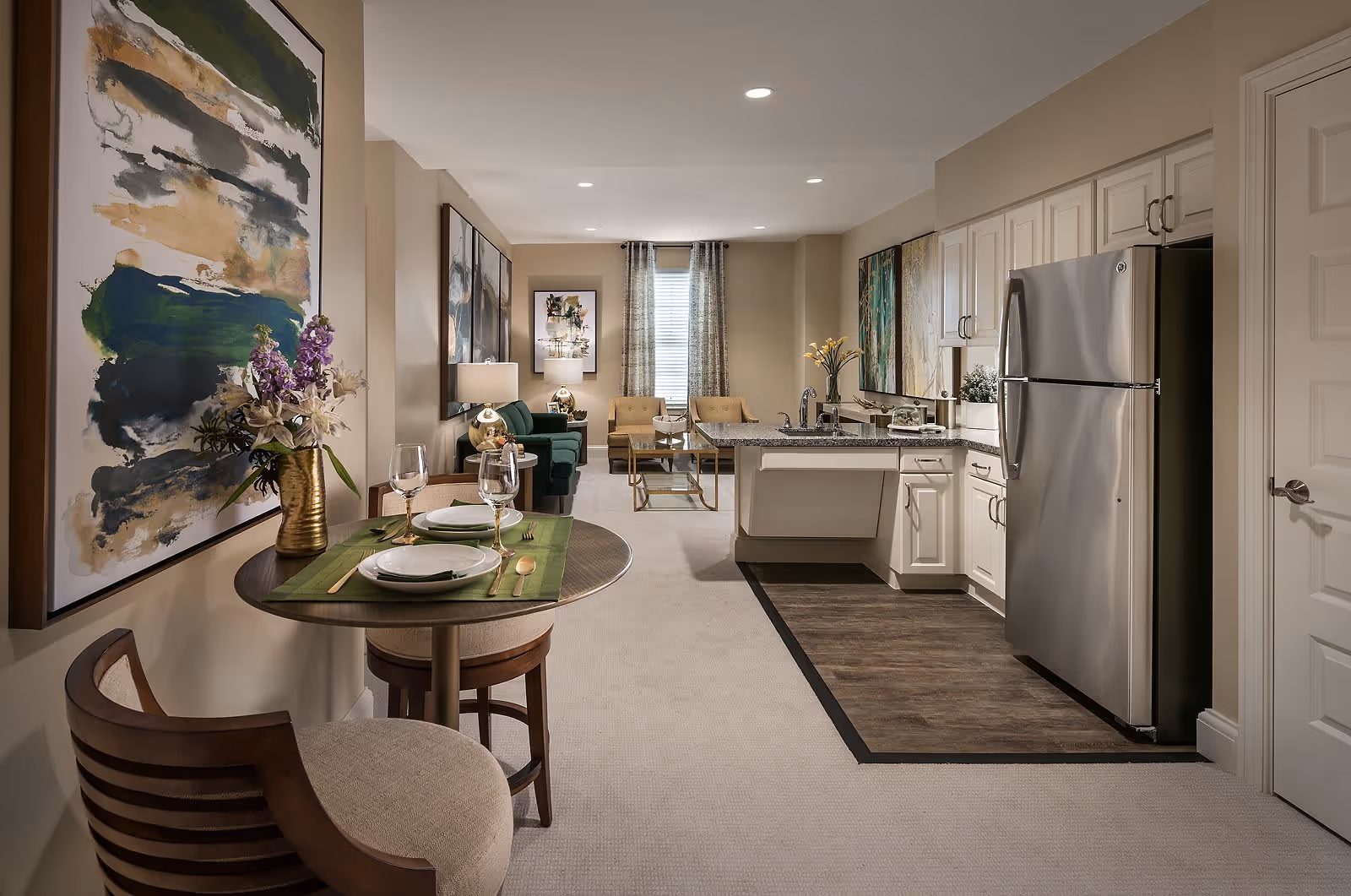 Interior view of a senior living facility apartment featuring a small dining area with a round table set for two, a kitchen with white cabinets and stainless steel refrigerator, and a living room with green and beige seating, decorative paintings on the walls, and a window with curtains in the background.