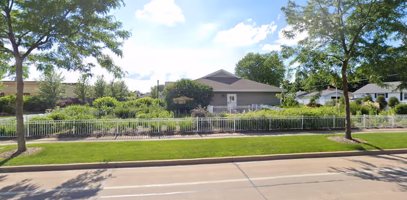 View of a single-story building surrounded by lush greenery and trees, with a white fence in front and a clear blue sky with some clouds above.
