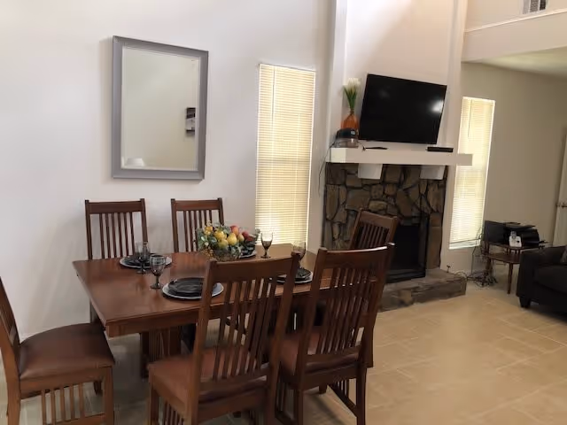 Dining area with a wooden table set for six with black plates and wine glasses. Behind the table is a stone fireplace with a flat-screen TV mounted above it. Two tall windows with blinds flank the fireplace. A mirror hangs on the wall to the left of the dining table. The room has beige tiled flooring and neutral-colored walls.