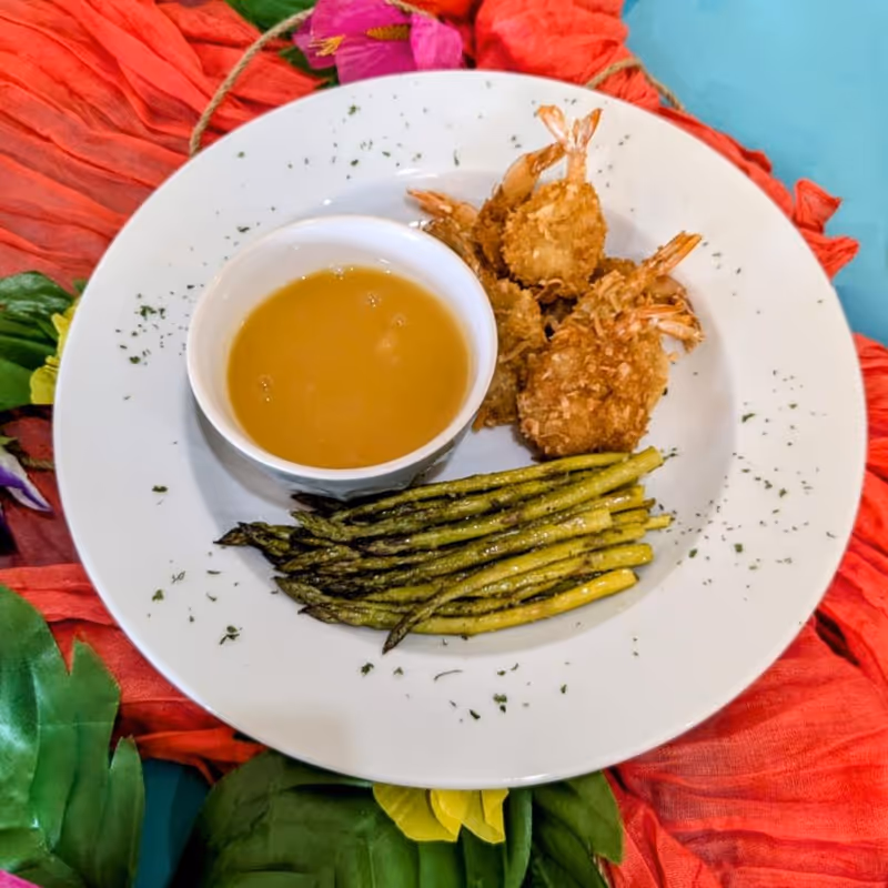 A white plate with breaded fried shrimp, grilled asparagus, and a small bowl of dipping sauce, placed on a colorful red and green floral tablecloth.