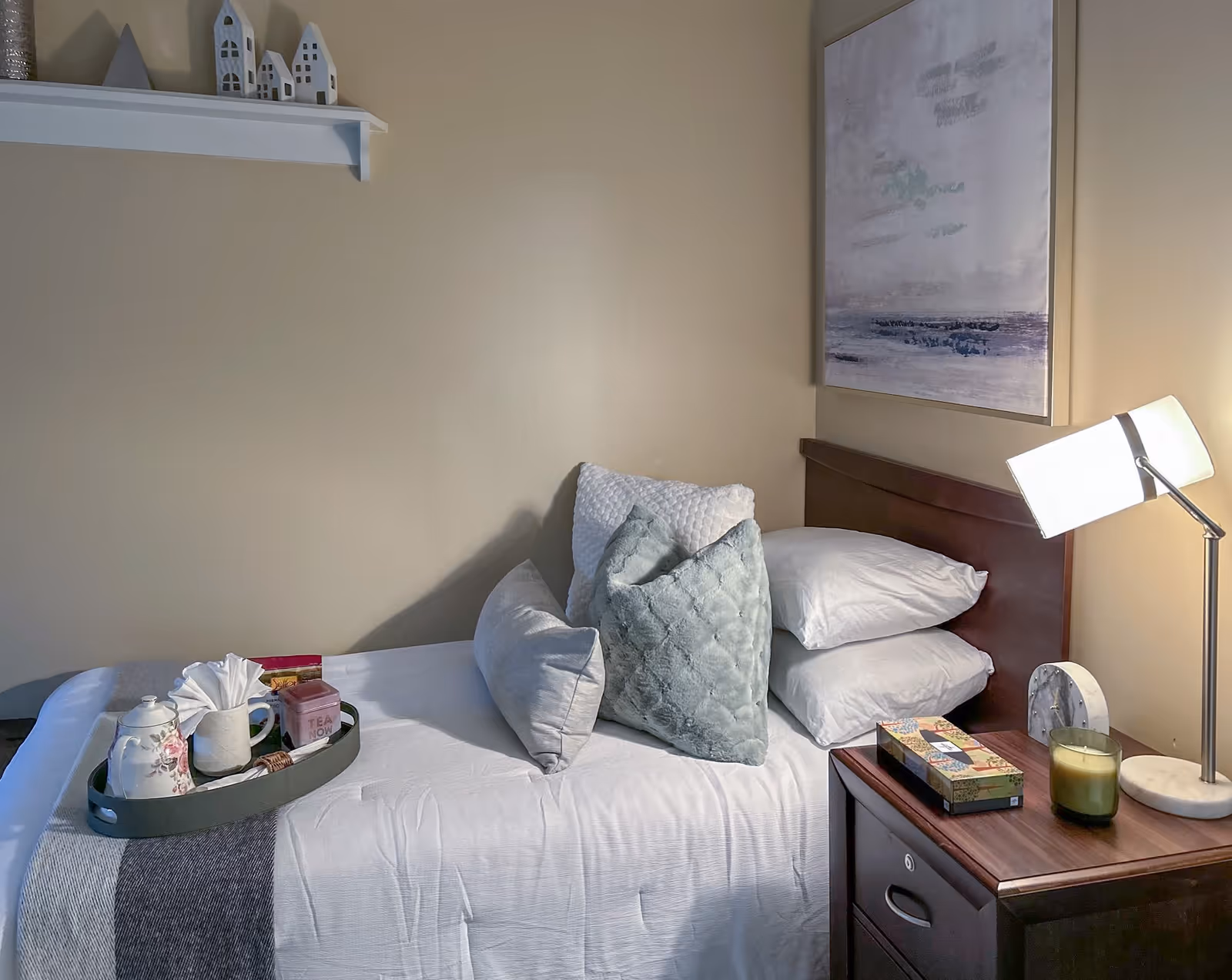 A neatly made single bed with multiple pillows and a tray holding a teapot, cup, and other items. Next to the bed is a wooden nightstand with a table lamp, a candle, and a decorative box. Above the bed is a framed abstract painting, and a small shelf with decorative house figurines is mounted on the wall.