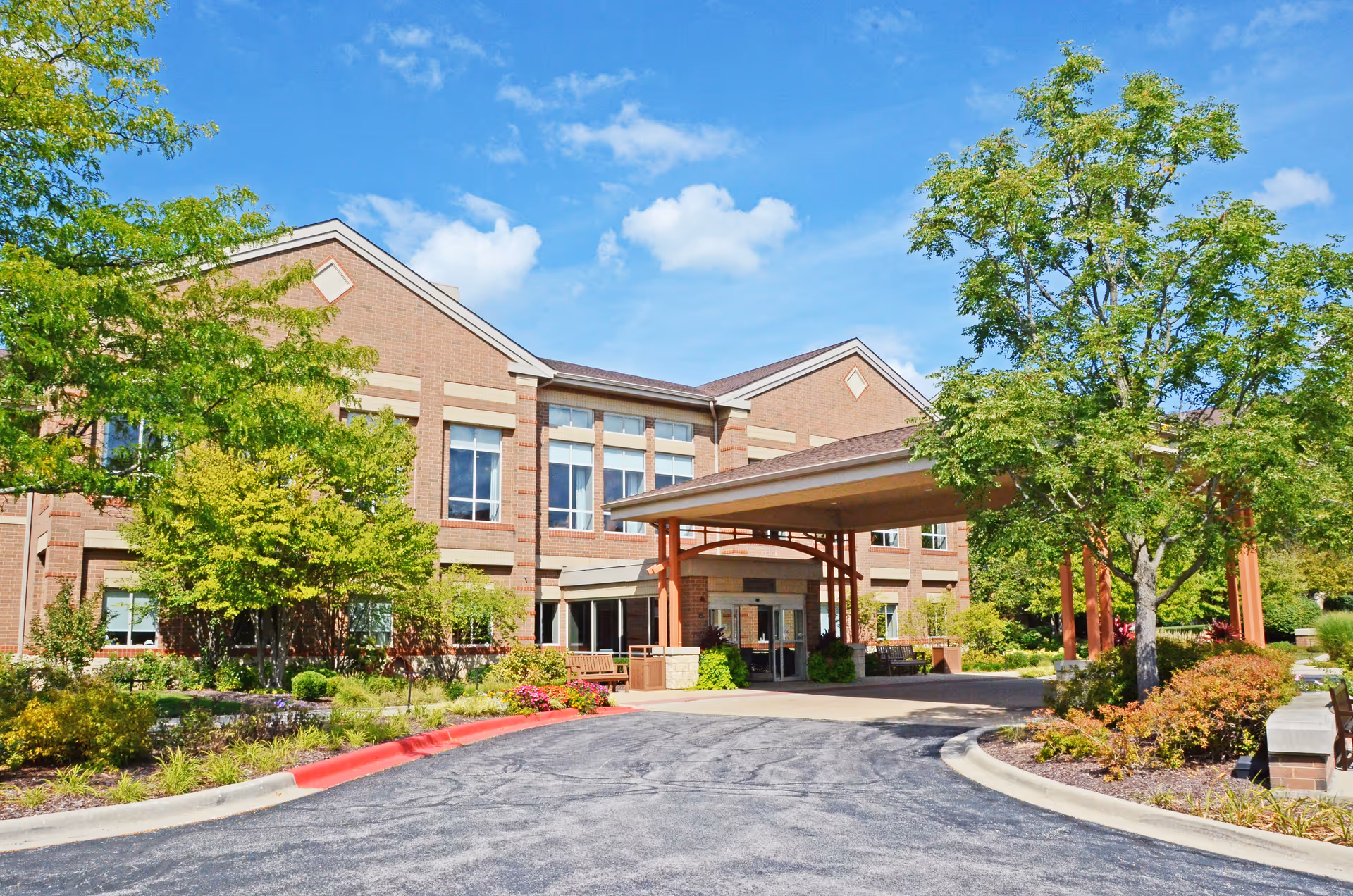Front exterior of a brick senior living building with a covered entrance, driveway, and landscaped trees and shrubs.