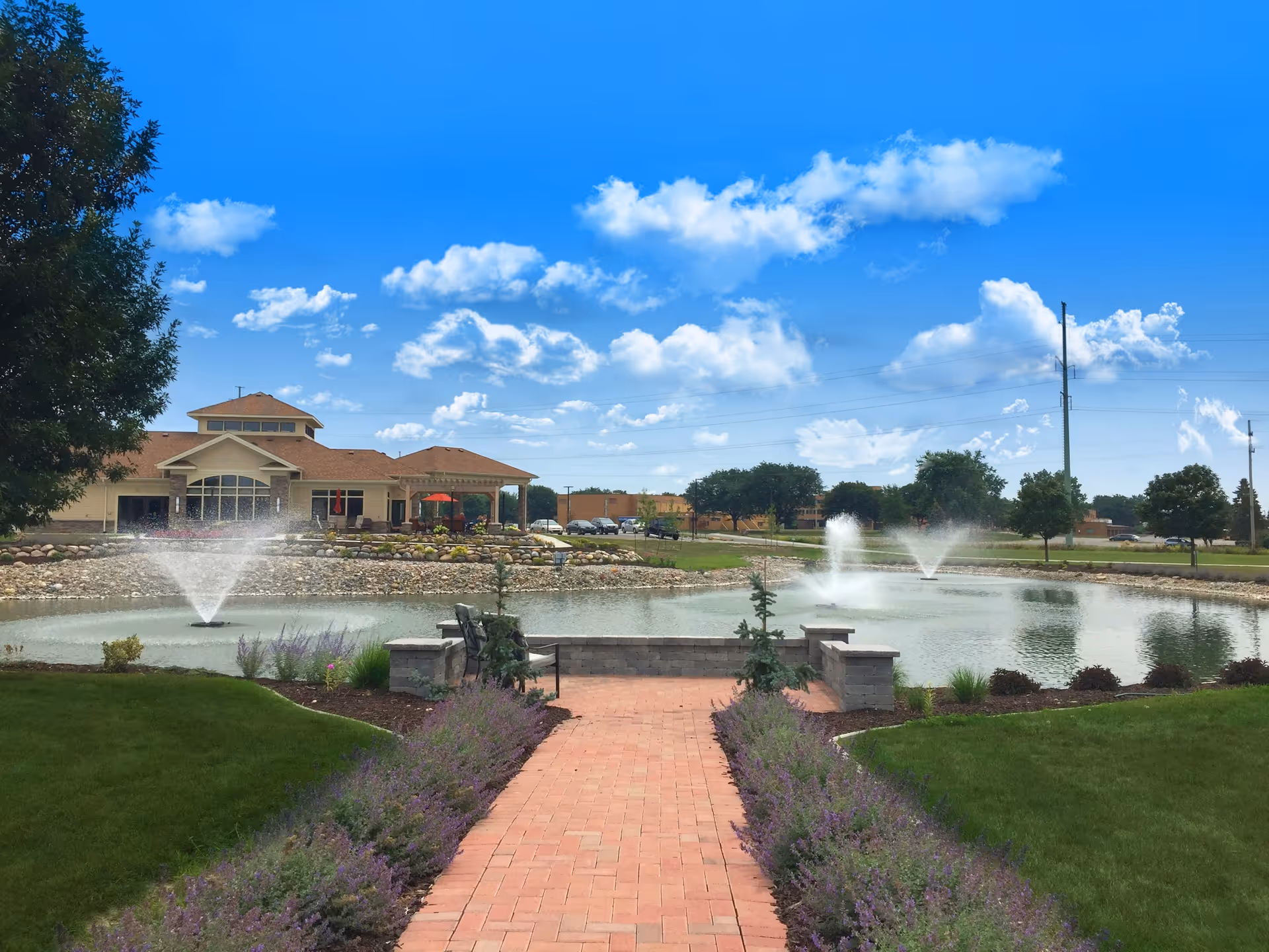 A scenic outdoor view of Wellspring Lutheran Services Senior Living Campus featuring a brick pathway lined with purple flowers leading to a small stone seating area by a pond with three water fountains. The building with a covered patio and red umbrellas is visible in the background under a bright blue sky with scattered clouds.