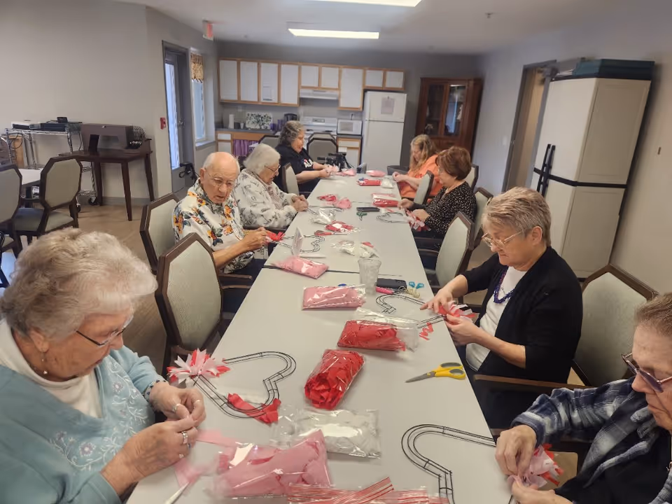 A group of elderly people sitting around a long table in a room with a kitchenette, engaged in a craft activity involving red and pink fabric pieces and wire frames shaped like hearts.