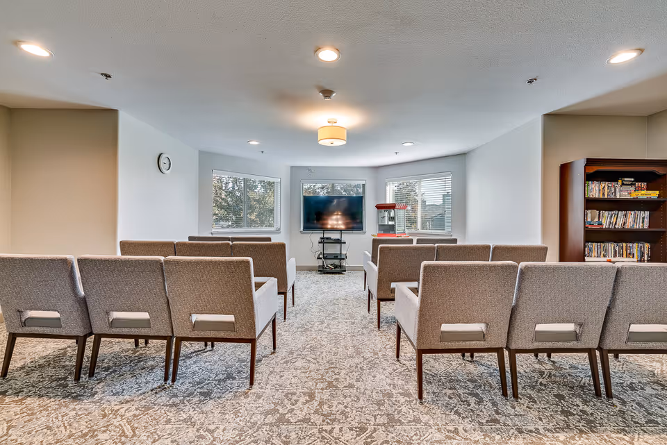A senior living facility common room with rows of gray upholstered chairs facing a flat-screen TV mounted on a stand. The room has three windows letting in natural light, a popcorn machine on a small table to the right of the TV, and a bookshelf filled with DVDs and books on the right side. The carpet has a patterned design, and the ceiling has recessed lighting and a central light fixture.