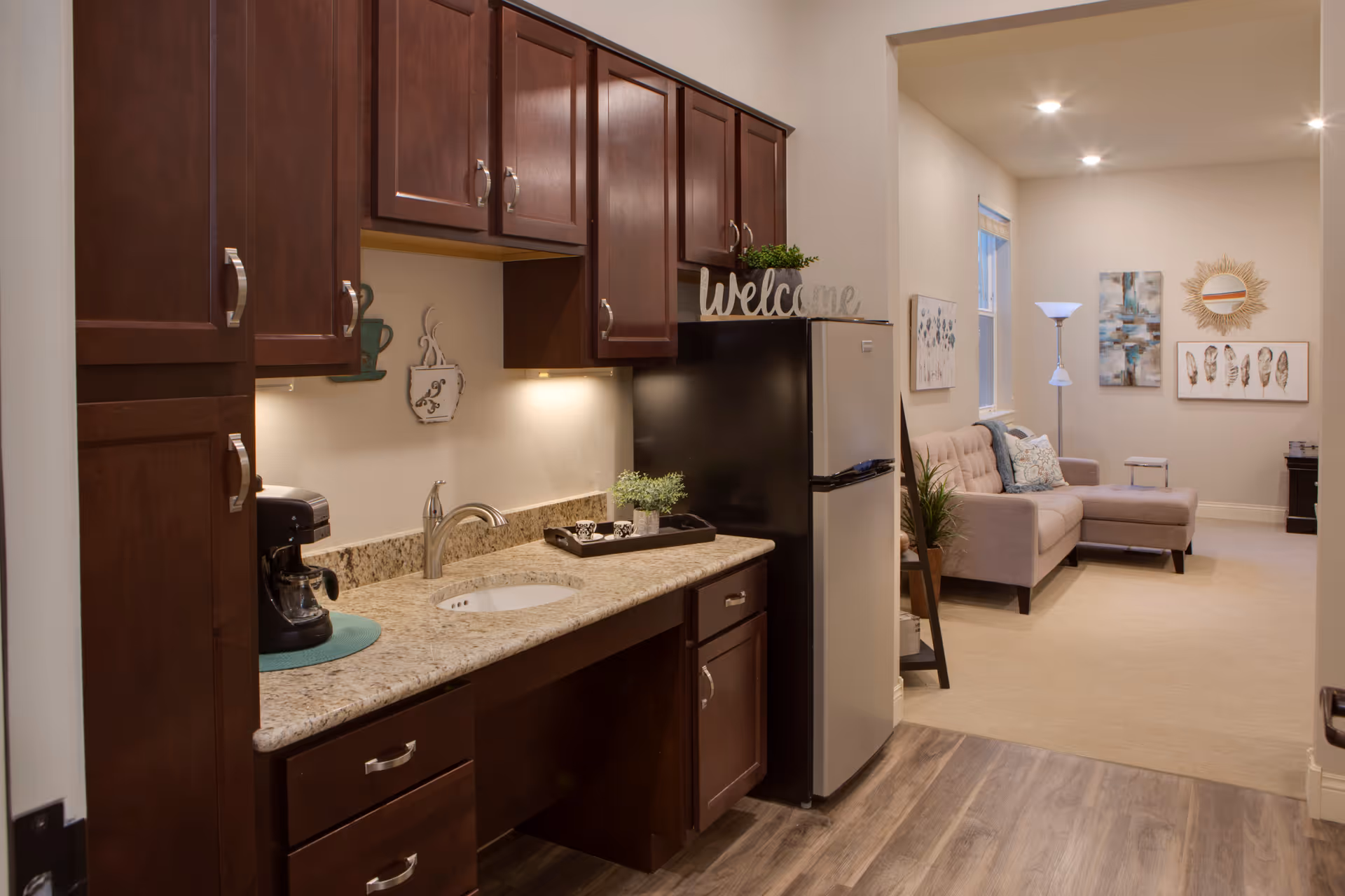 Interior view of a senior living facility unit showing a kitchenette with dark wood cabinets, a granite countertop, a coffee maker, and a small sink. Next to the kitchenette is a stainless steel refrigerator with a decorative 'welcome' sign on top. In the background, there is a living area with a beige sectional sofa, a floor lamp, wall art, and a small side table.