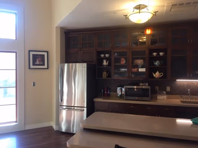 Interior view of a kitchen area featuring dark wooden cabinets with glass doors displaying teapots and cups, a stainless steel refrigerator, a microwave on the countertop, a sink, and a two-tiered kitchen island with a light fixture above.