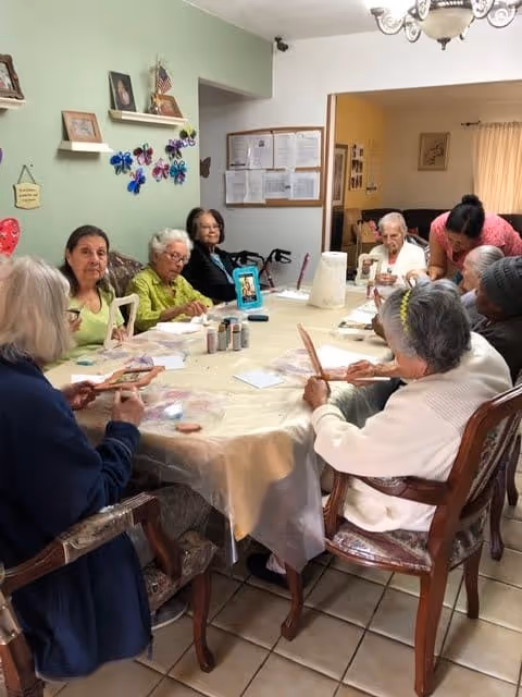 A group of elderly women sitting around a large table in a well-lit room, engaging in a craft activity. The table is covered with a plastic tablecloth and various craft supplies. The room has light green and white walls with framed pictures and decorative items. A caregiver is assisting one of the women.