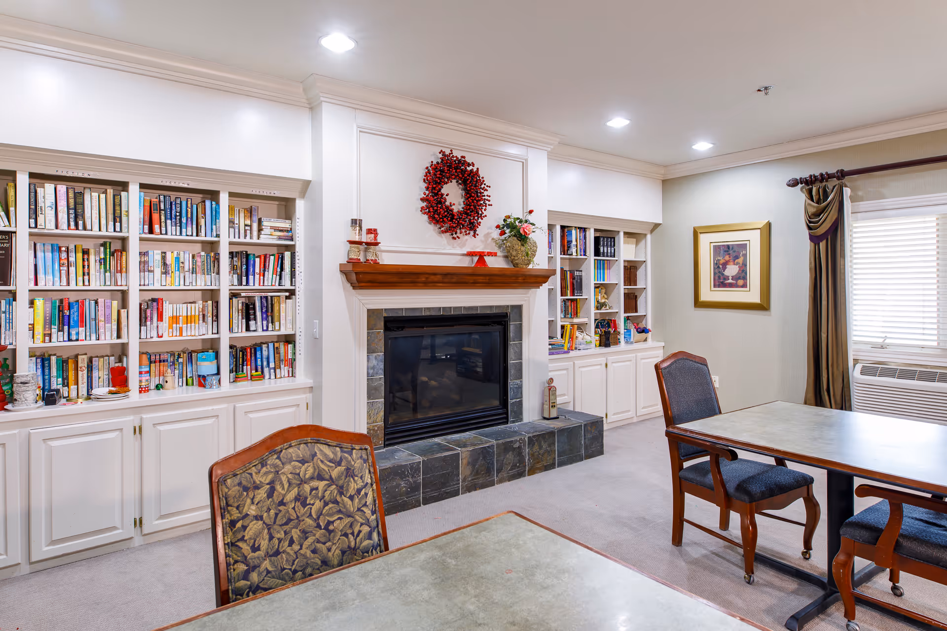 A cozy interior room featuring a fireplace with a red wreath above it, flanked by built-in white bookshelves filled with books and decorative items. There are tables and chairs with patterned upholstery, a window with blinds and curtains, and a framed picture on the wall.