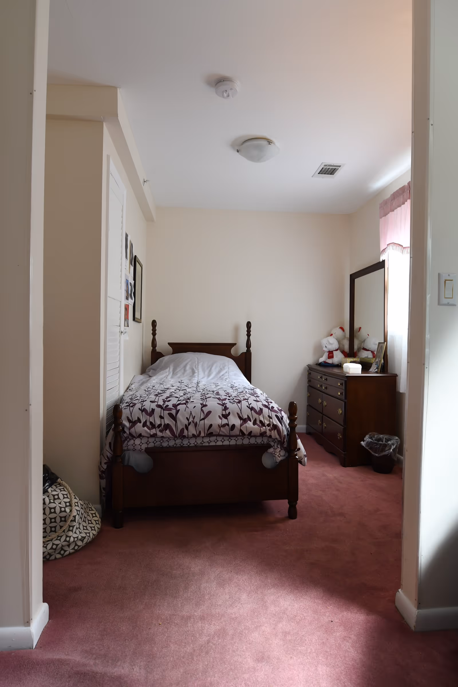 Small bedroom with a single wooden bed, dresser and mirror, pink carpet and stuffed animals on the dresser.