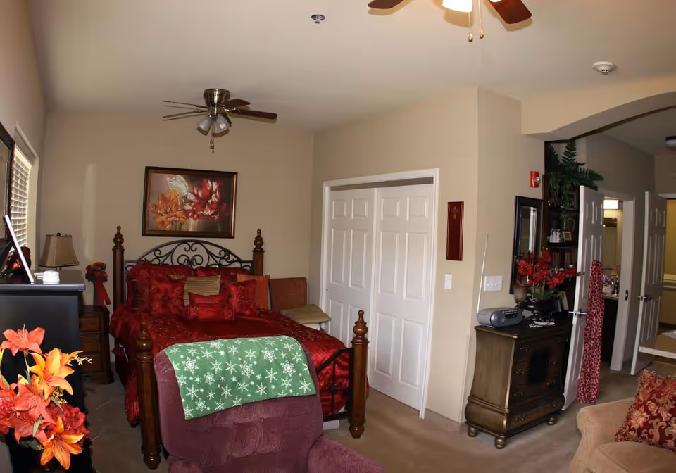A cozy bedroom in a senior living facility featuring a bed with red bedding and multiple pillows, a ceiling fan with lights, a wooden dresser with a mirror and decorative items, a maroon armchair with a green blanket, and floral decorations. There is a closet with white sliding doors and an open doorway leading to another room.