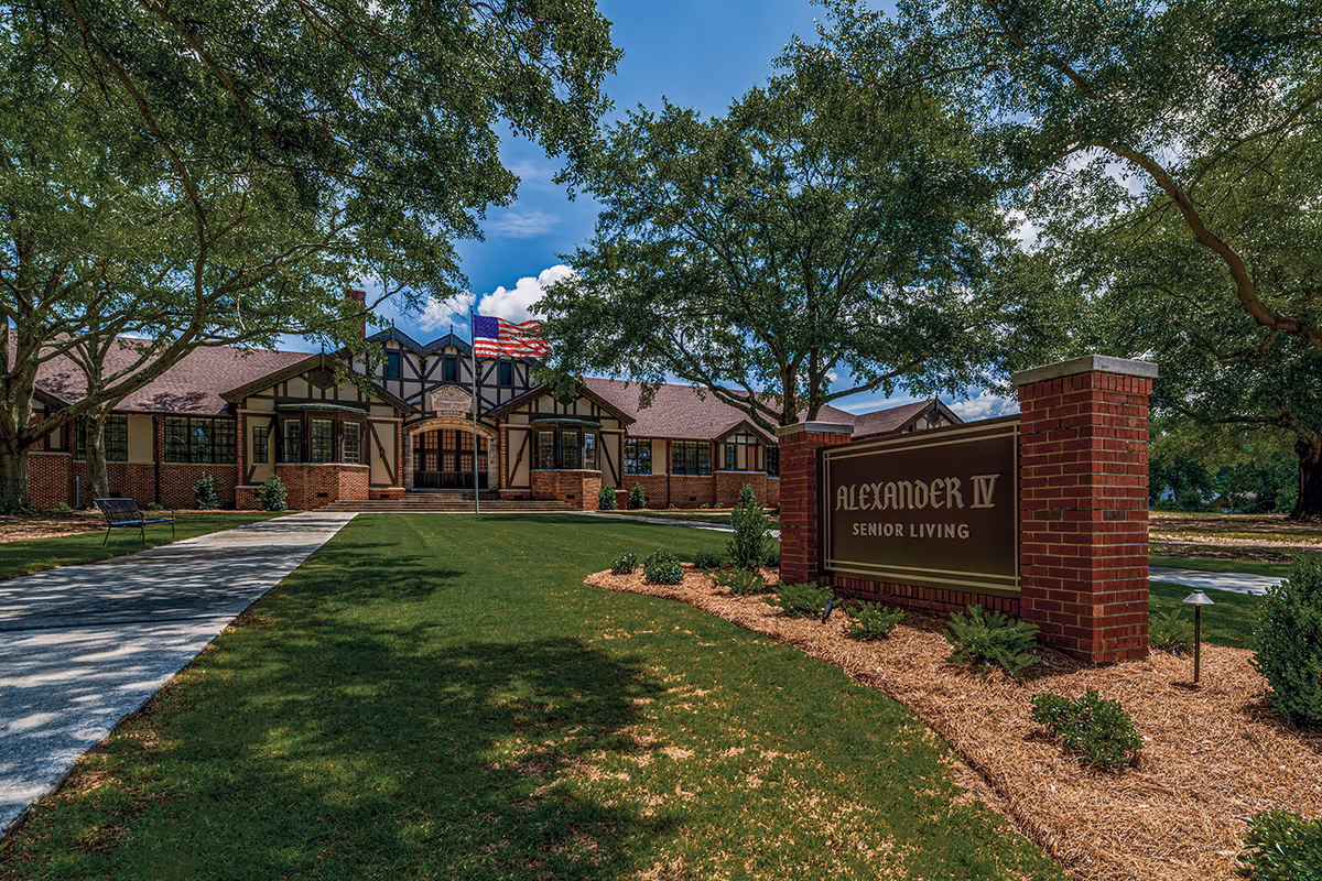 Exterior view of Alexander IV Senior Living facility with a brick and wood building, an American flag on a flagpole, a paved walkway, green lawn, trees, and a large sign with the facility name.