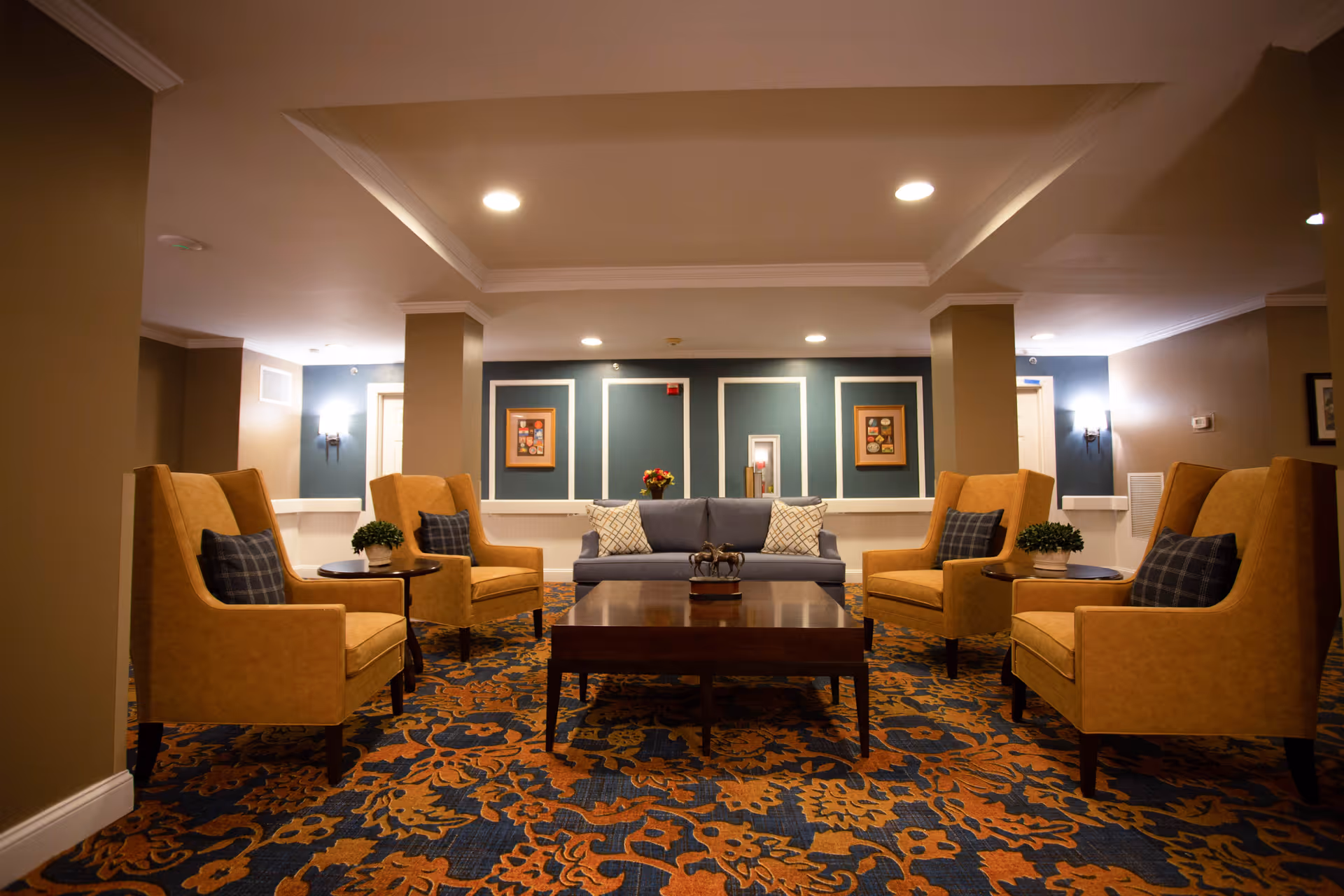 A cozy living room area with a blue sofa and four mustard yellow armchairs arranged around a dark wooden coffee table. The room features a patterned carpet in blue and orange tones, beige walls, and framed artwork on the far wall. There are small plants on side tables next to the armchairs and recessed ceiling lights providing warm illumination.