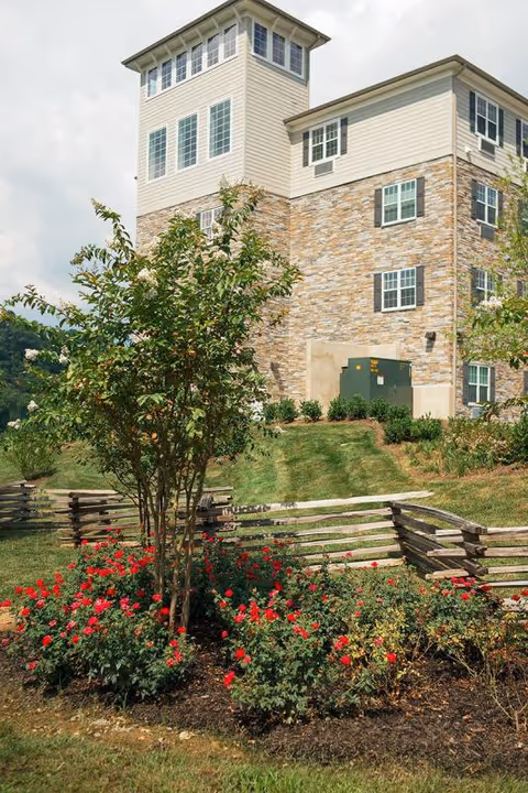 A landscaped garden area with red flowering bushes and a small tree in front of a multi-story building with stone and beige siding. The building has multiple windows and a fenced area with wooden rails. The sky is partly cloudy.
