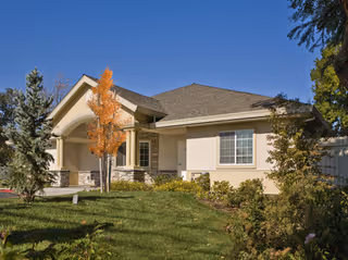 Single-story building with beige exterior walls, a gray shingled roof, and a small covered porch. The front yard has green grass, bushes, and a tree with orange leaves. The sky is clear and blue.