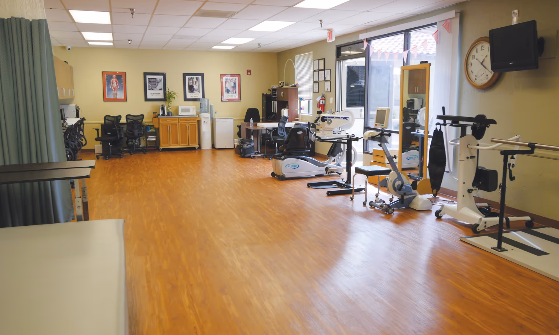 Spacious rehabilitation/exercise room with wood-look flooring, medical exercise equipment, chairs and a wall clock.