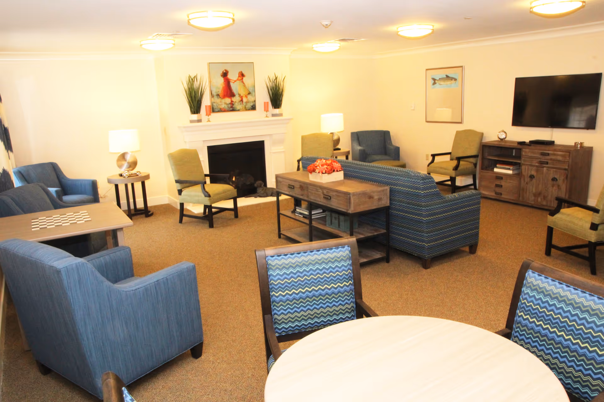 A cozy senior living common area with multiple armchairs and sofas arranged around a fireplace. There is a wooden coffee table with a flower arrangement in the center, a TV mounted on the wall, and framed artwork above the fireplace and on the wall. The room is warmly lit with ceiling lights and table lamps.