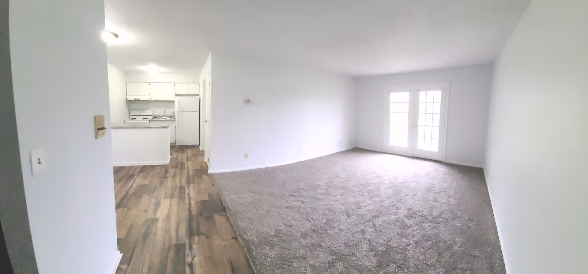 An empty interior space featuring a living room area with carpet flooring and a kitchen area with wood-style flooring. The living room has white walls and a set of French doors letting in natural light. The kitchen includes white cabinets, a refrigerator, a stove, and a countertop.