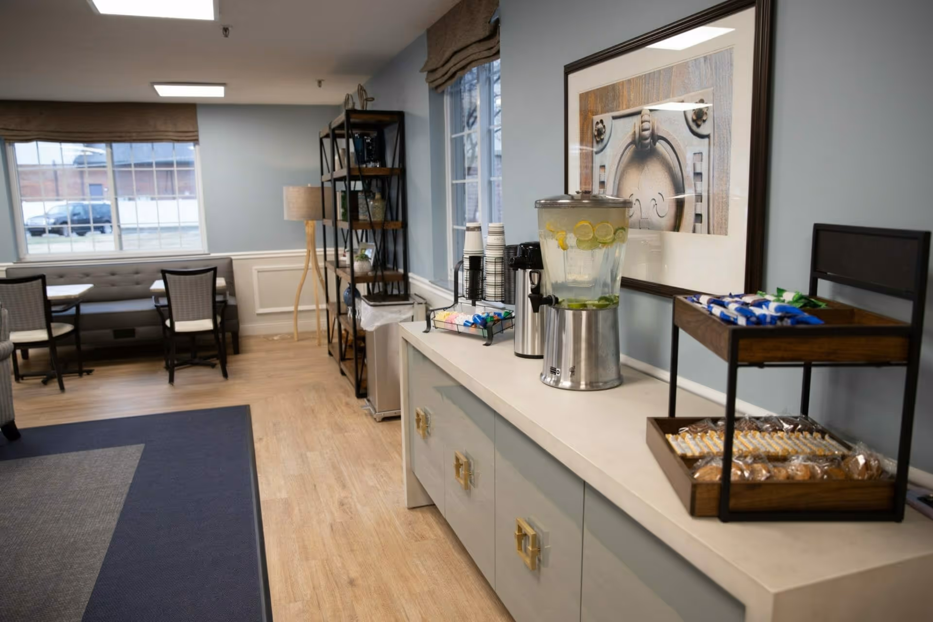 Communal interior room with seating, tables, a shelving unit, and a sideboard holding a beverage dispenser and snacks.