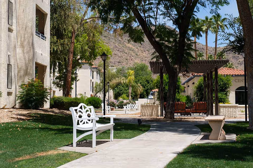Outdoor pathway in a senior living facility with white benches along the walkway, trees providing shade, a pergola with seating underneath, and buildings in the background with a mountainous landscape.