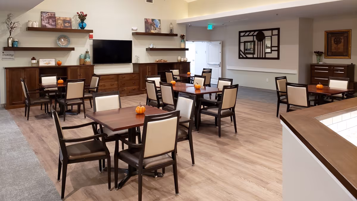 A dining area in an assisted living facility with several wooden tables and chairs arranged neatly. Each table has a small decorative pumpkin on it. The room features wooden flooring, a wall-mounted flat-screen TV, and shelves with decorative items such as vases and framed pictures. The walls are painted in neutral tones, and there is an exit door visible in the background.
