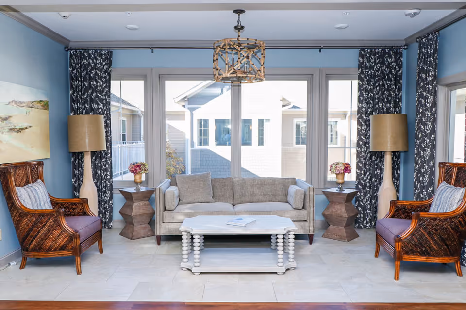A bright living room with a beige sofa centered between two dark wooden armchairs with purple cushions. There are two matching wooden side tables with flower vases on each side of the sofa, flanked by tall beige lamps. Large windows with dark patterned curtains let in natural light, and a white coffee table with a book on it sits in front of the sofa. A decorative light fixture hangs from the ceiling.