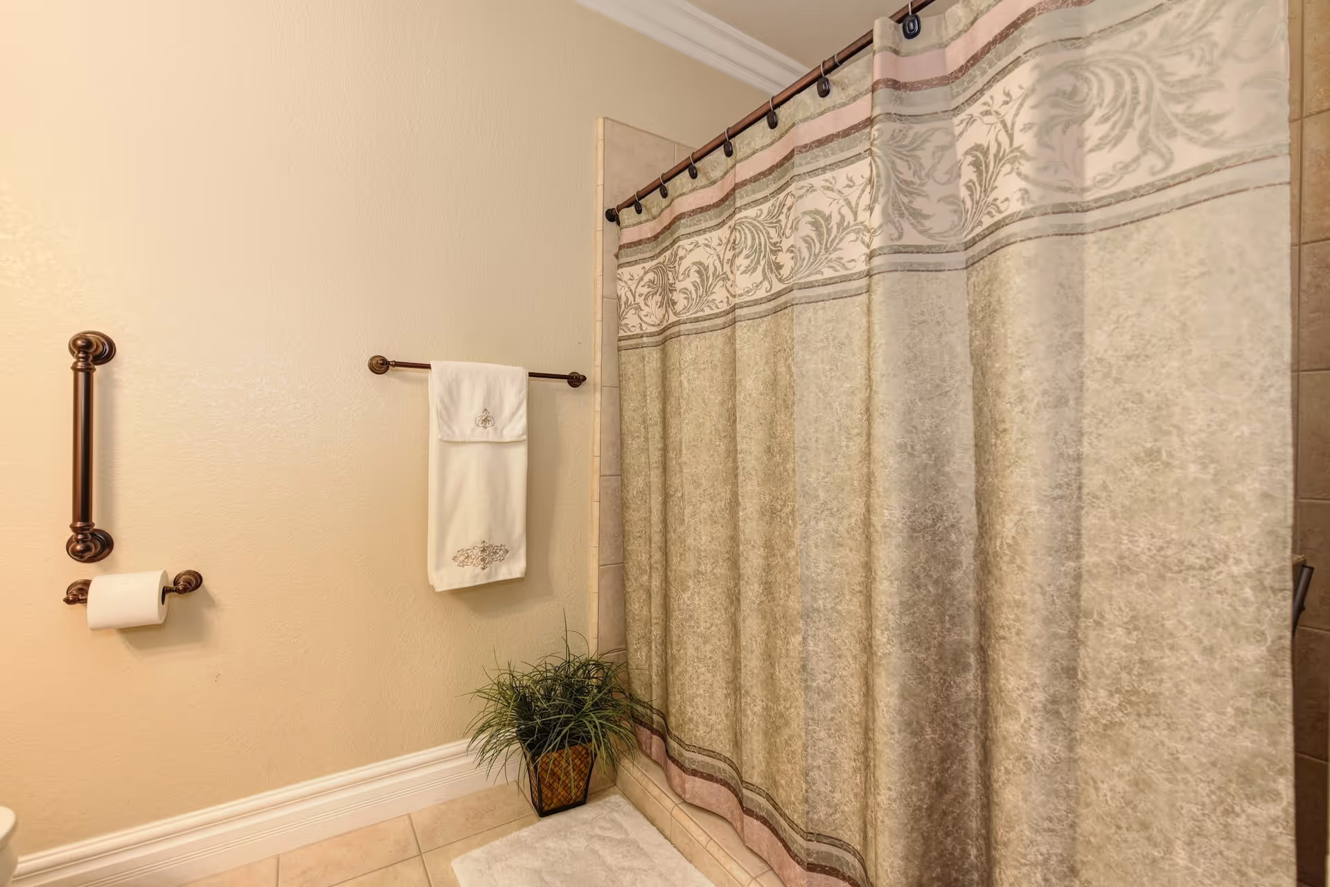 A bathroom with a beige and brown patterned shower curtain, a white towel hanging on a bronze towel rack, a toilet paper holder with a roll of toilet paper, a bronze grab bar, a small green potted plant on the floor, and beige tiled flooring.