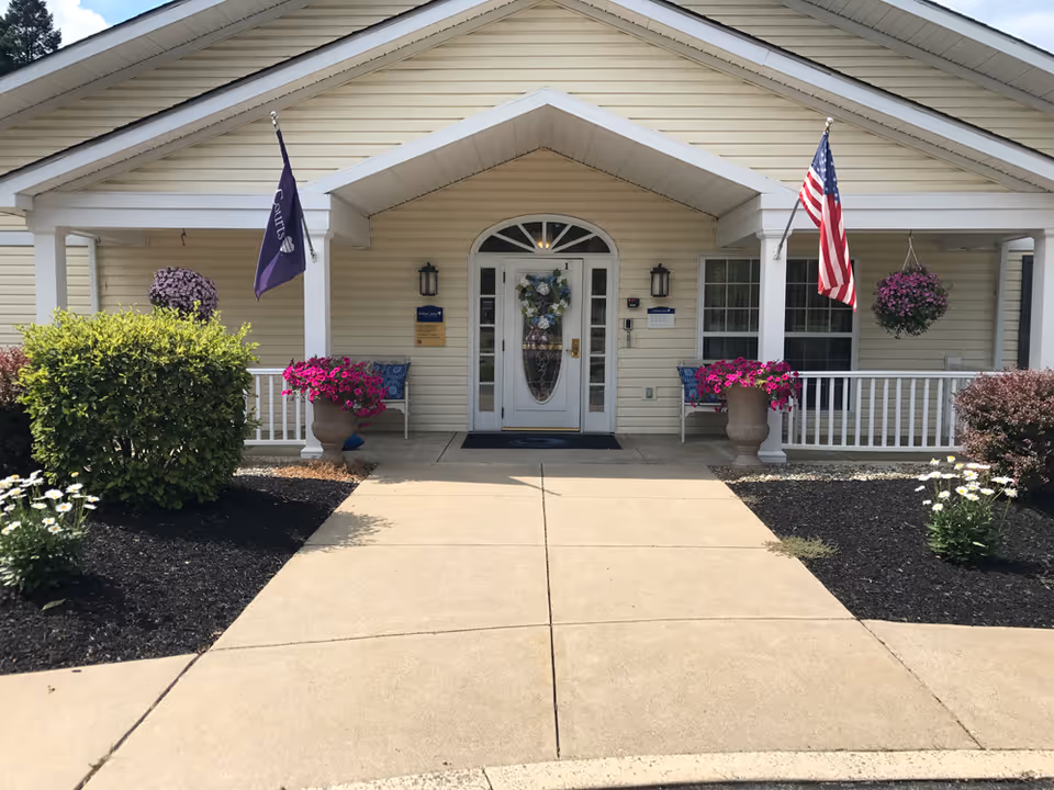 Front entrance of Arden Courts - ProMedica Memory Care Community in Allentown, showing a beige building with a white door decorated with a floral wreath. There are two flags on either side of the entrance, one American flag and one purple flag. The entrance is flanked by flower pots with bright pink flowers and benches with cushions. The walkway leading to the door is concrete, bordered by landscaped bushes and flowers.