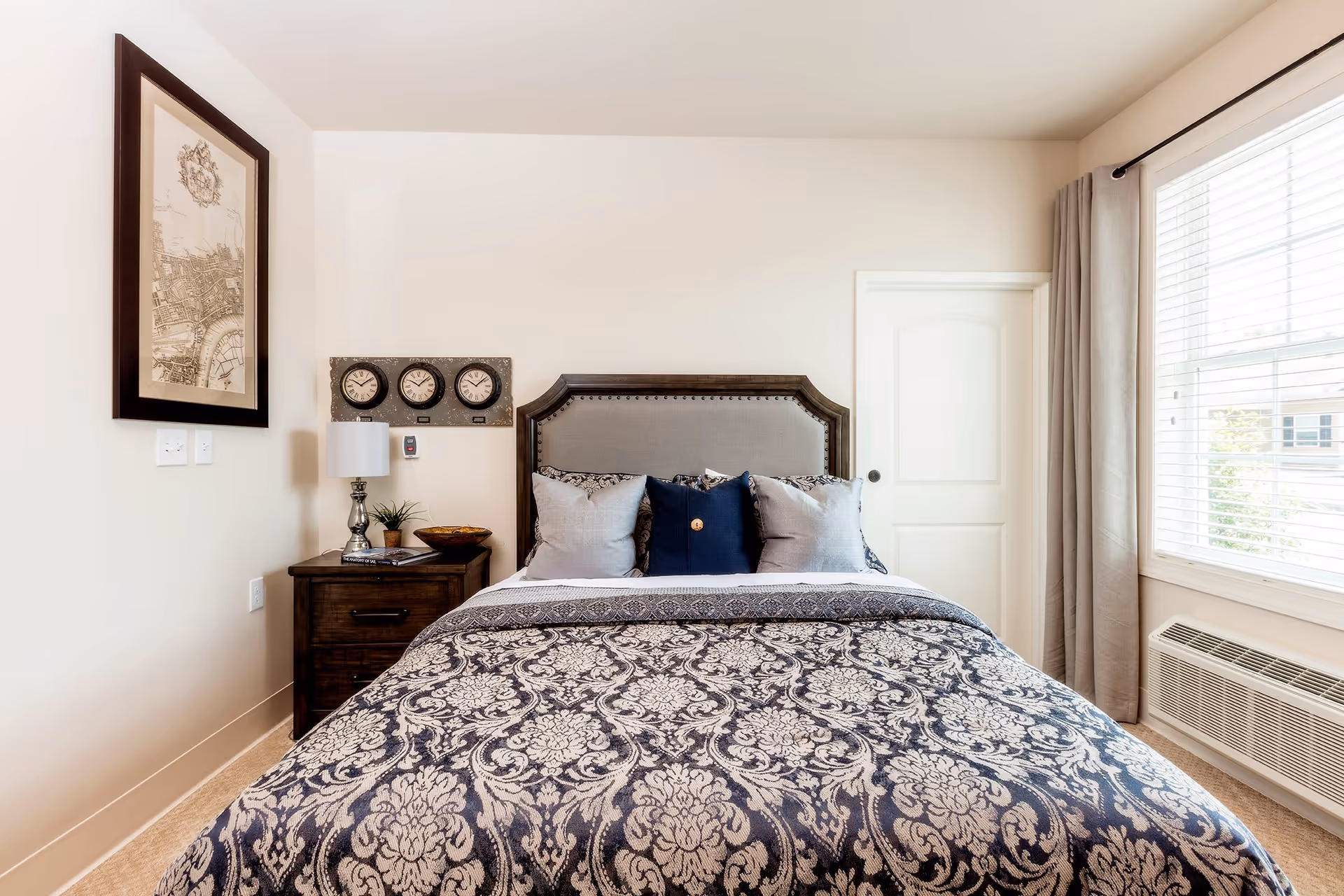 A neatly made bed with a patterned dark blue and white bedspread and multiple pillows in a bright bedroom. There is a wooden nightstand with a lamp, a small plant, and a bowl next to the bed. Above the nightstand are three clocks showing different times and a framed map on the wall. A window with blinds and curtains is on the right side, letting in natural light.
