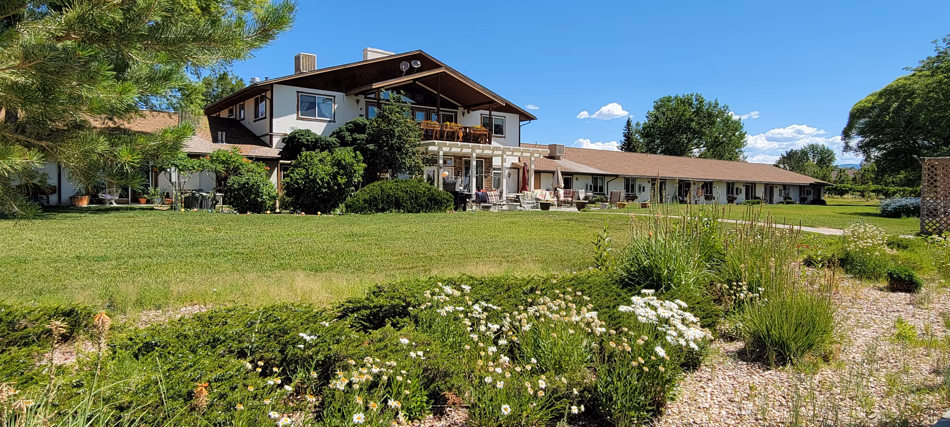 A large senior living facility building with a two-story section and a long single-story wing, surrounded by green lawns, bushes, and flowering plants under a blue sky with scattered clouds.