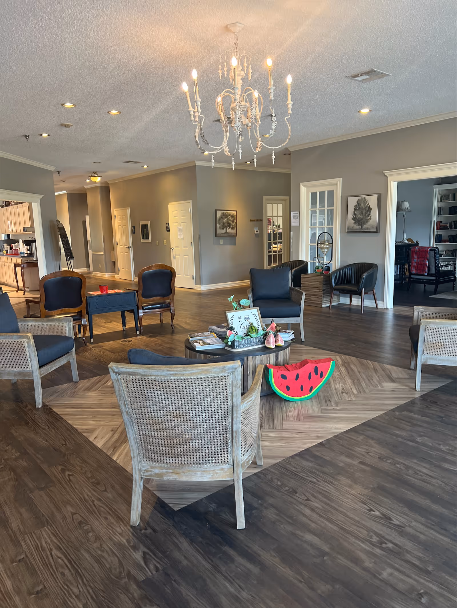 A spacious and well-lit common area in a senior living facility named Arbor Rose, featuring a chandelier, several chairs arranged around a circular coffee table with magazines and decorative items, and a watermelon-shaped cushion on the floor. The room has wood-patterned flooring, gray walls, and doorways leading to other rooms.