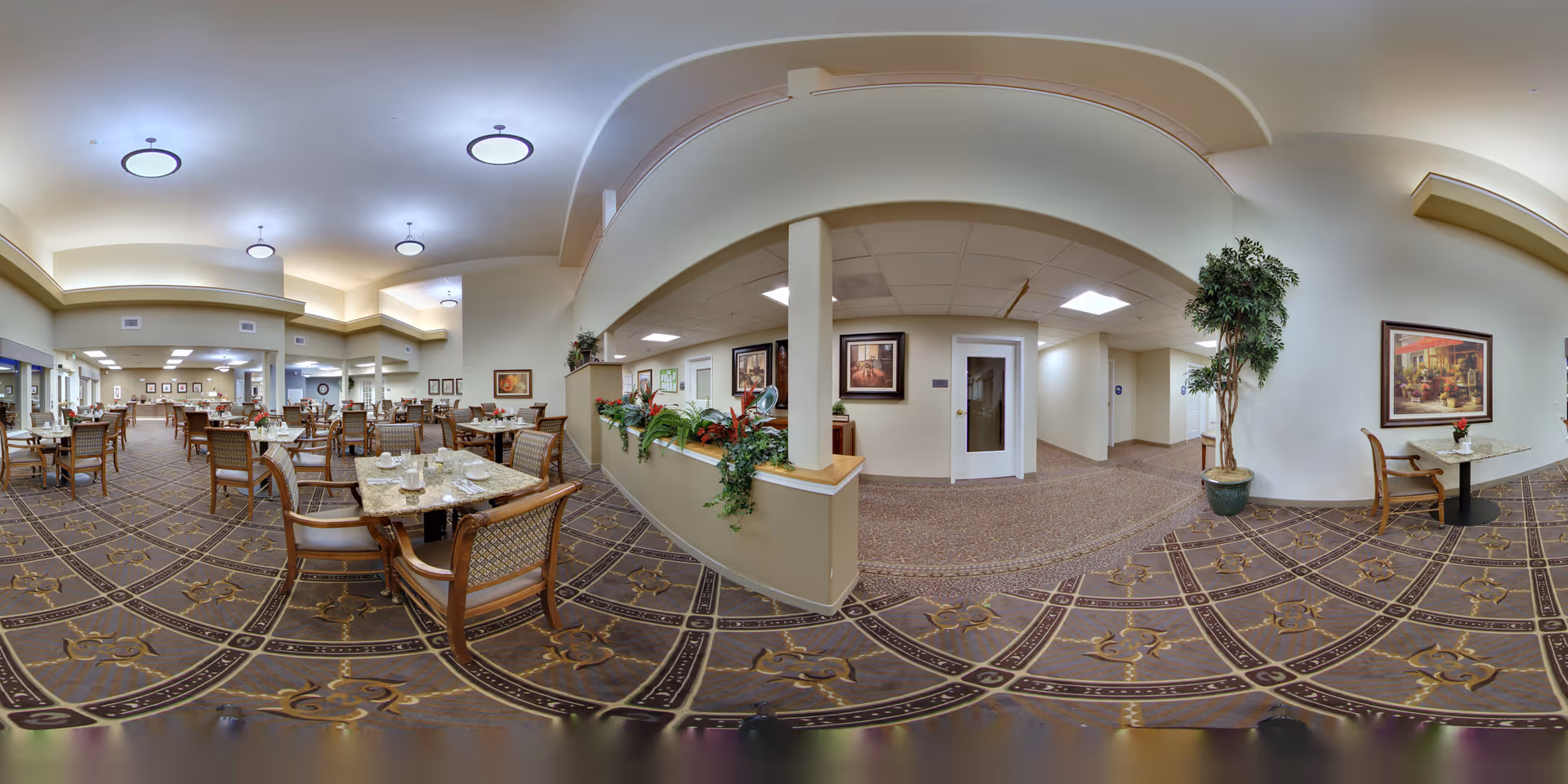 Wide view of a spacious dining room in a senior living facility with multiple tables and chairs arranged neatly. The room features patterned carpet, soft lighting from ceiling fixtures, framed artwork on the walls, and some greenery including potted plants and a planter with flowers. There is a small seating area with a table and chairs near the entrance hallway.