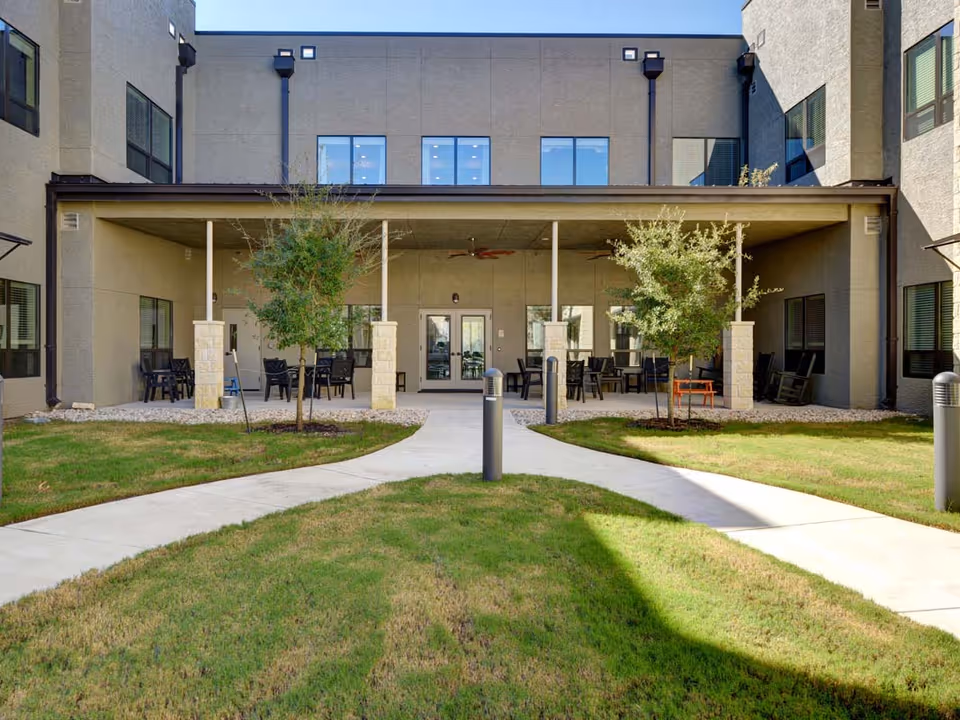 Outdoor courtyard area of a senior living facility with a covered patio featuring multiple tables and chairs. The patio is supported by stone pillars and has ceiling fans. There are two small trees planted in grassy areas with concrete walkways leading to the patio entrance. The building has multiple windows and a beige exterior.