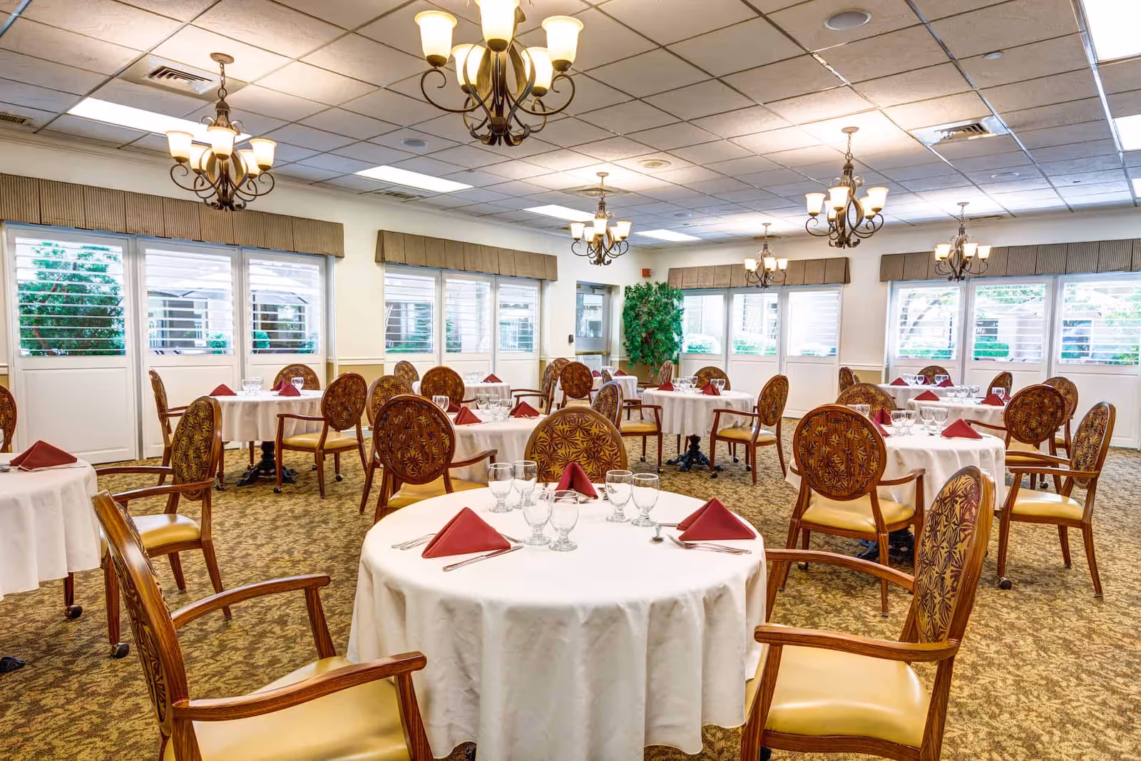 Bright dining room with round tables set with white tablecloths, folded red napkins and ornate chairs under chandeliers.