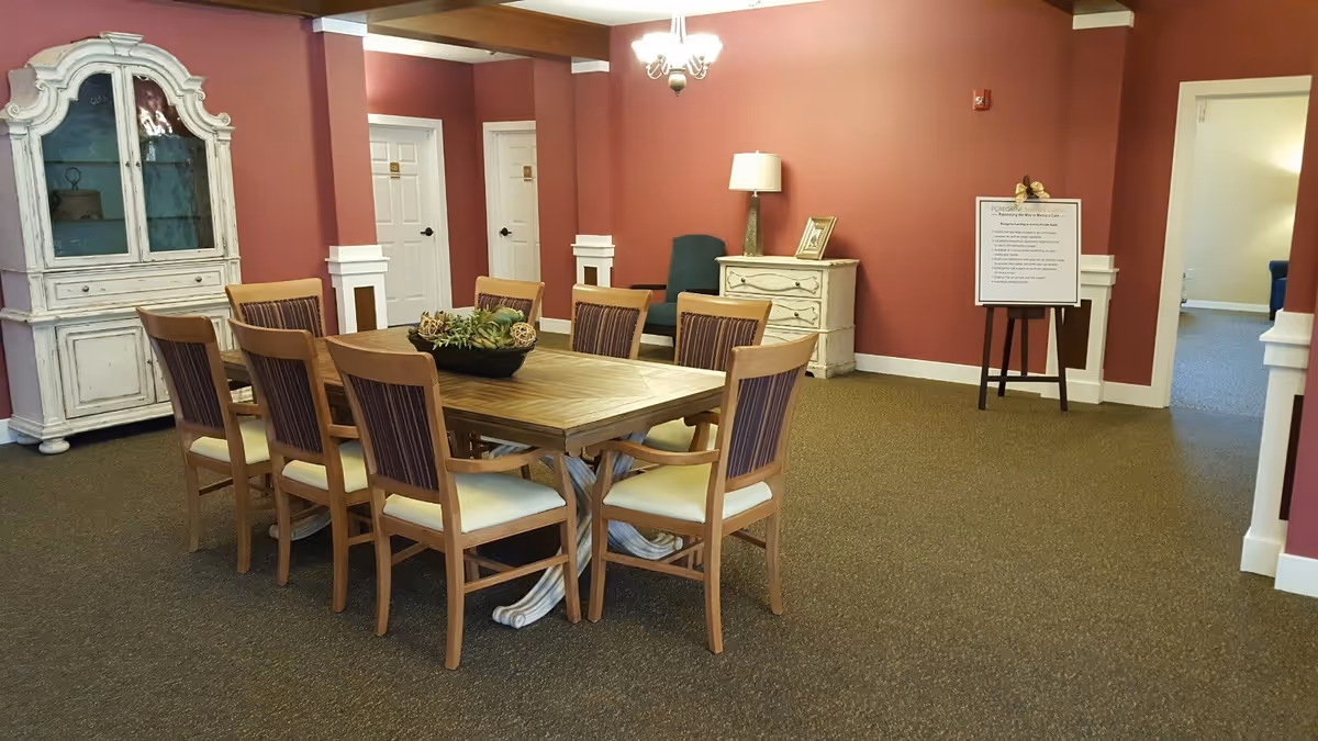 A dining room with a wooden table surrounded by eight chairs with cushioned seats and striped backs. The walls are painted a deep red color with white trim. There is a white vintage-style cabinet on the left side and a white dresser with a lamp and picture frame on top against the far wall. A green upholstered chair is placed next to the dresser. A chandelier hangs from the ceiling, and a sign on an easel is positioned near an open doorway on the right.
