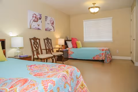 Two pastel beds and seating area fill a well-lit senior living bedroom.