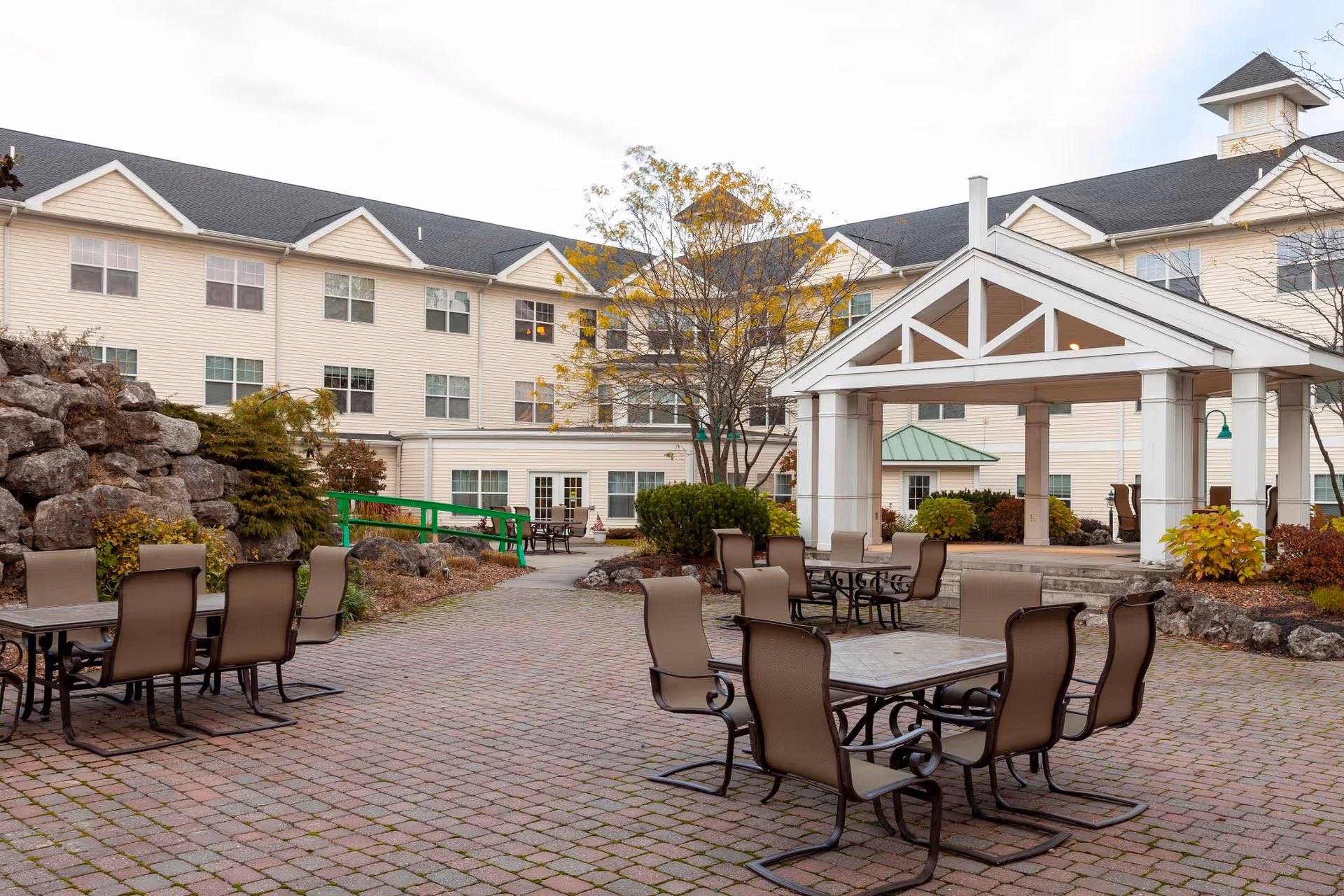Outdoor courtyard area of a senior living facility with multiple tables and chairs arranged on a brick-paved surface. The courtyard features a white pavilion structure with a peaked roof and columns. Surrounding the courtyard are landscaped areas with rocks, bushes, and trees, and a multi-story beige building with many windows in the background.