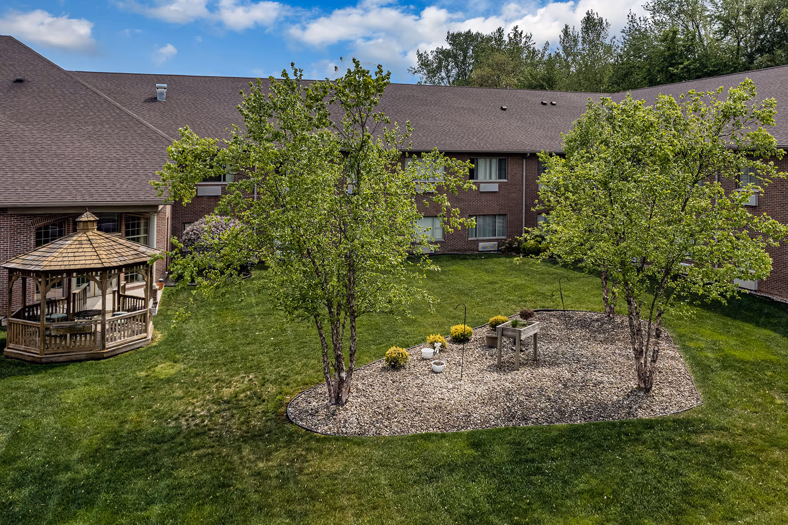Outdoor courtyard area of Brentwood at Elkhart Independent Living featuring a green lawn, two leafy trees planted in a rock-covered garden bed, a wooden gazebo with seating, and a brick building with multiple windows in the background under a partly cloudy sky.