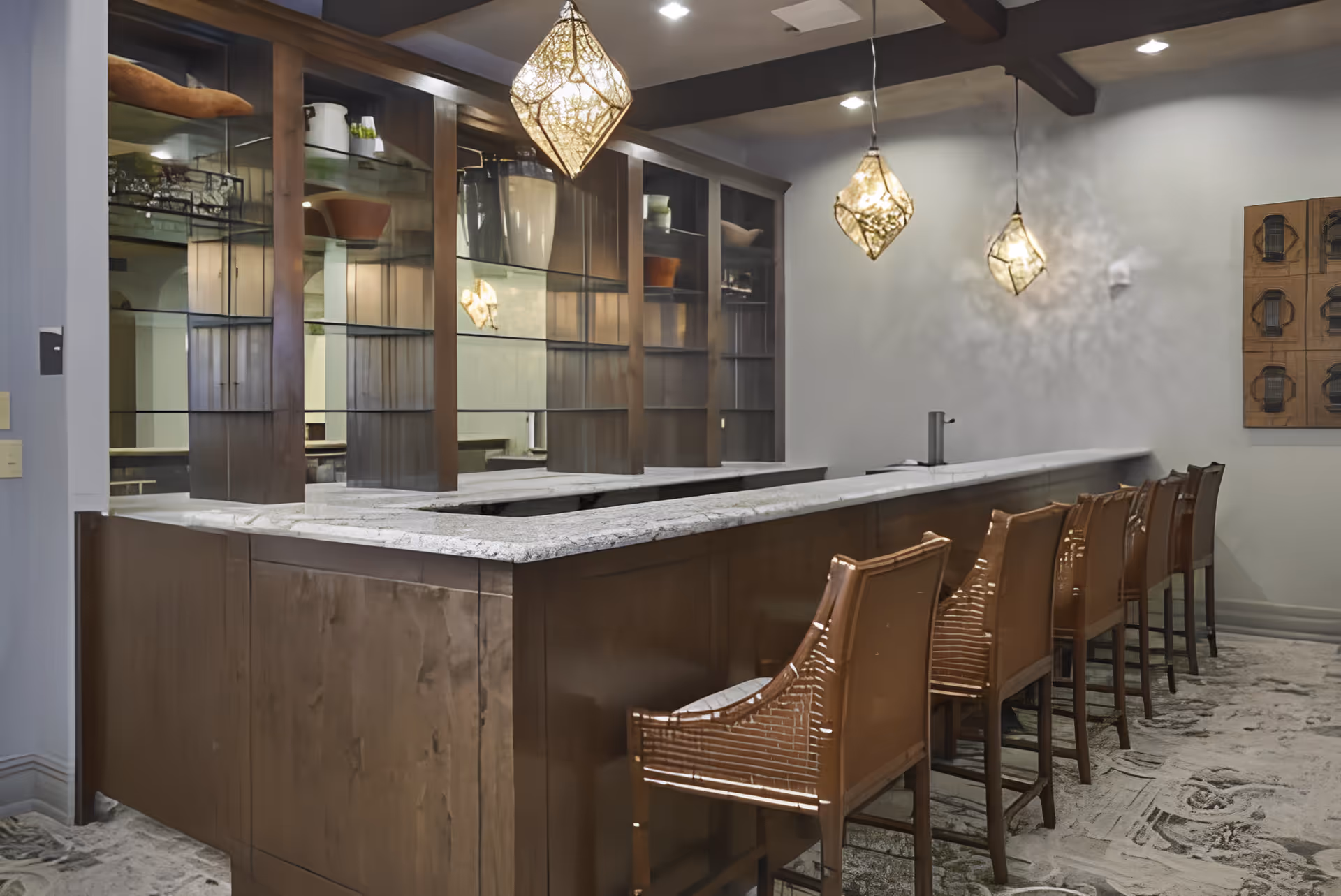 Interior view of a bar area with a long marble countertop and six wooden bar stools. Above the counter hang three decorative pendant lights with intricate designs. Behind the bar is a wooden shelving unit with glass shelves and various decorative items. The floor is carpeted with a patterned design, and the walls are painted light gray with a piece of wall art visible on the right side.