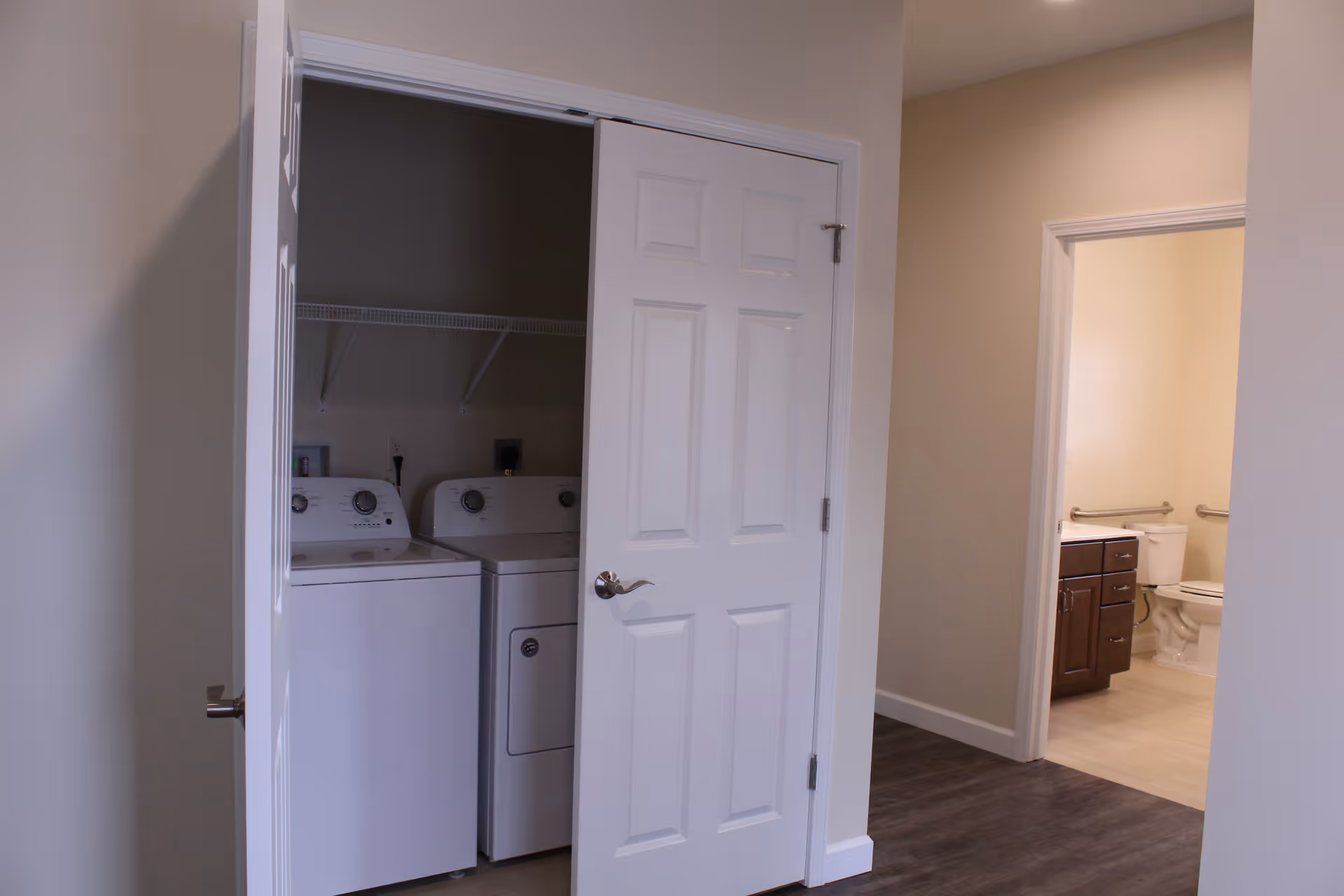 Interior view showing a laundry area with a washing machine and dryer behind a partially open white door. To the right, there is a bathroom with a toilet, wooden vanity with drawers, and grab bars on the walls.
